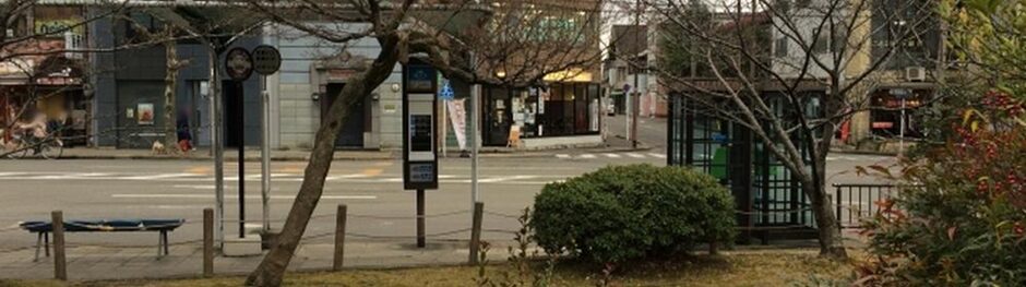 街路樹や植え込み越しに見える住宅街と交差点、公衆電話ボックスが映る落ち着いた街並みの風景