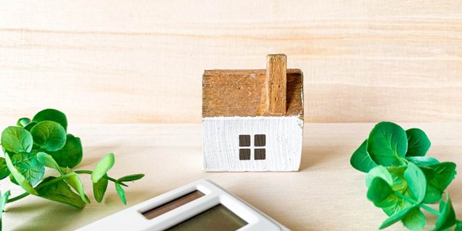 木製の小さな家と電卓、周囲に緑の葉が添えられた省エネのイメージ